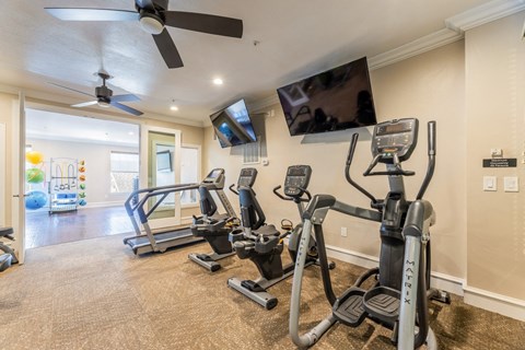 Cardio machines including an elliptical, cycling machines, and a treadmill facing a wall-mounted tv in the fitness center at Encore at Buckingham apartments in Richardson, TX